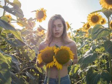 20 'li yaşlarda, güzel, çıplak, uzun saçlı, gülümseyen ve göğsünü güneş batarken sararmış ayçiçeği tutan bir kadın fotoğrafı..