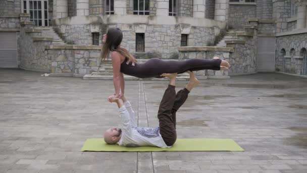 Ajuste pareja deportiva practicando acro yoga con pareja juntos en la esterilla al aire libre. Pareja haciendo ejercicio acrobático. Acróbata hembra se equilibra en las piernas de su pareja masculina — Vídeo de stock