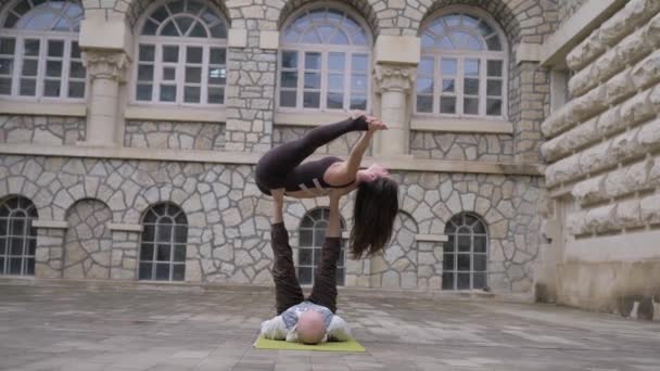 杂技瑜伽。一个女人靠在她的背上，靠在男人的腿上保持平衡，在加洛根加做一个分开的练习。室外 — 图库视频影像