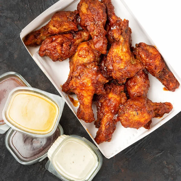 Takeaway box of crispy grilled chicken legs with a spicy marinade served with a hot chili dipping sauce, on a dark stone surface with copyspace. 1:1