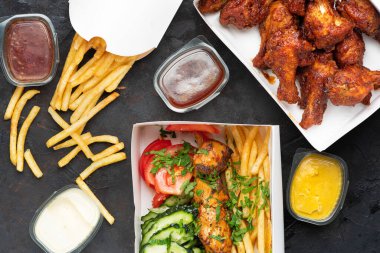 Fast food: Bir kutu kızarmış tavuk budu, Yunan souvlaki 'si ve fotokopi uzayı olan koyu taştan bir yüzey üzerinde patates kızartması. Fast food teslimatı. Menü için üst görünüm Yakın plan