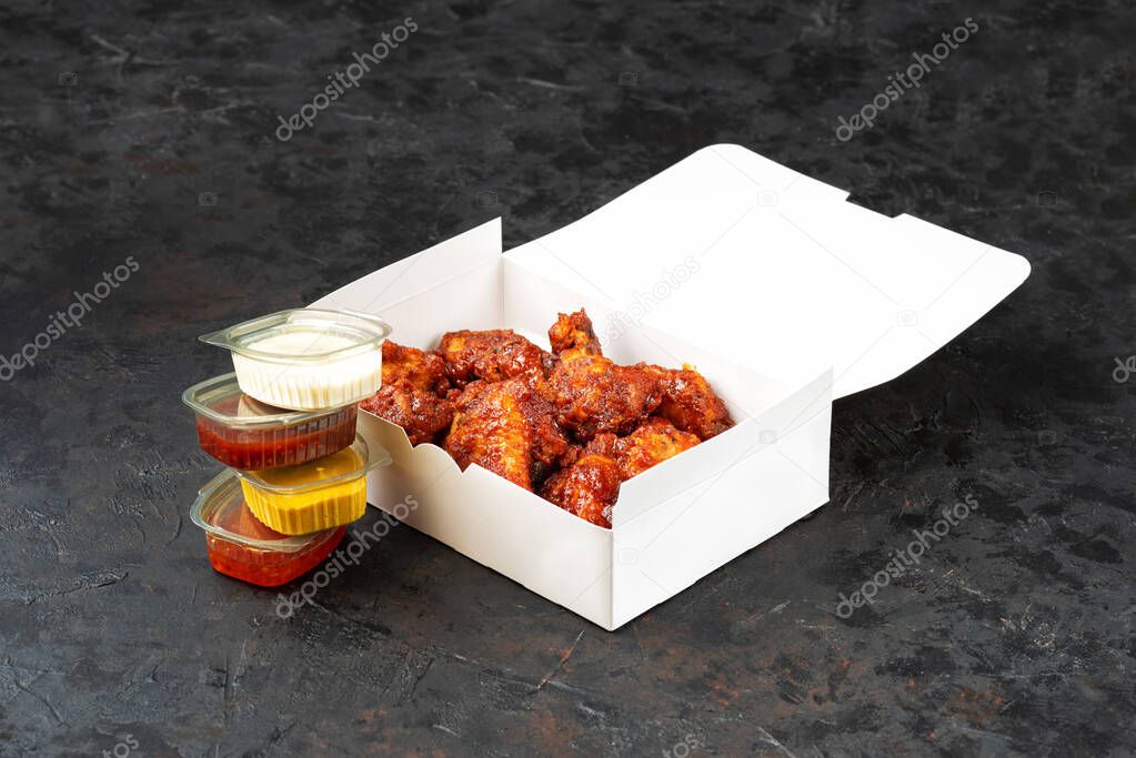 Takeaway box of crispy grilled chicken legs with a spicy marinade served with a hot chili dipping sauce, on a dark stone surface with copyspace.