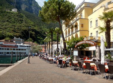 Riva del Garda, İtalya - Mayıs 2017: Göl manzaralı restoranlar ve arka plandaki düzenli feribotlardan biri ile gezinti alanı boyunca görünüm