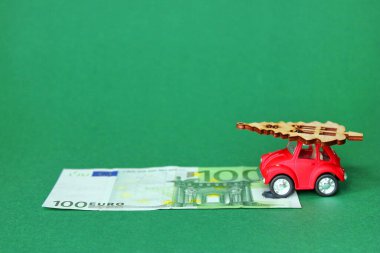 100 Euro ve Noel ağacı taşıyan kırmızı bir oyuncak araba. Çatıda Noel ağacı ahşap figürlü küçük kırmızı bir oyuncak araba. Önünde yeşil arka planda 100 Euro var..