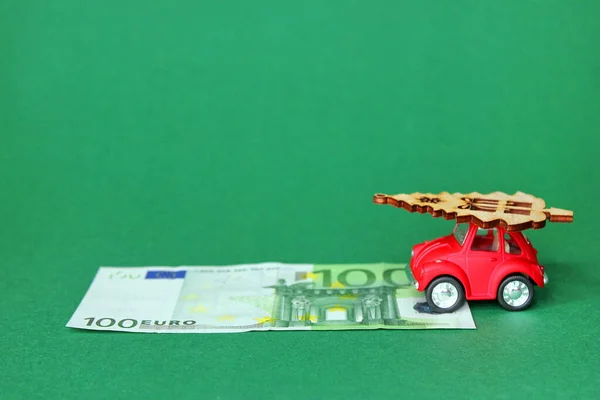 100 Euros and a red toy car carrying a Christmas tree. A small red toy car with a wooden figure of a Christmas tree on the roof. A 100 Euro bill in front of her on a green background.