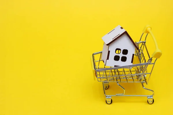 黄色の背景にスーパーマーケットのカートで小さな木造の家。プロパティを購入するアイデア。大きな買い物 — ストック写真