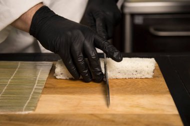 Suşi ustası Japon rulosunu tahtadan bıçakla kesti. Şef siyah koruyucu eldivenlerle yemek hazırlıyor. Pirince sarılmış suşi rulosu. Restoran mutfağında profesyonel pişirme işlemi.