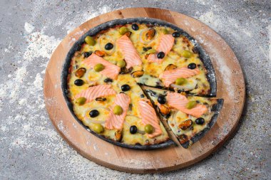 Ahşap tabakta deniz mahsulü siyah hamurlu pizza. Mutfak unu ve gri beton arka planda baharatlarla servis ediliyor. Fast food Dish. Somon dilimleri, yeşil ve siyah zeytin ve midye.