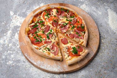 Salam dilimleri, domates, mozzarella peyniri ve taze yeşilliklerle dolu tam pizza. Ahşap tabakta servis edilir. Mutfak unu ve baharatlar gri beton arka planda servis edilir..