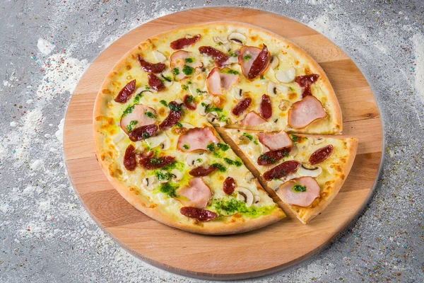 Whole rustic Pizza with smoked hunting sausage, ham, mushrooms, mozzarella cheese served with pesto sauce on wooden plate served with culinary flour and spices on gray concrete background.