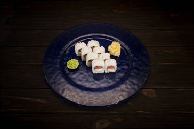Japon suşi rulosu, wasabi sosu ve koyu mavi cam tabakta zencefil ve ahşap zemin ile servis edilir. Çiğ somon fileto, pirinçli krem peynir ve yosun. Suşinin içi dışına çıktı.