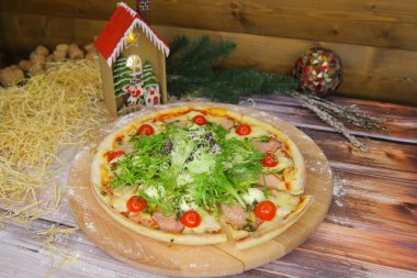 Tahta tabakta servis edilen İtalyan pizzası ve arka planda Noel süsü olan bir masa. Taze marul, roka, tavuk, jambon dilimleri, parmesan peyniri, bıldırcın yumurtası, vişne domatesli fast food yemeği.