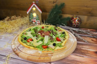 İtalyan pizzası ahşap tabakta ve masada servis edilir ve arka planda yeni yıl süsü vardır. Taze marul, jambon, jambon, jambon, parmesan peyniri, zeytin, vişne domatesli fast food yemeği.