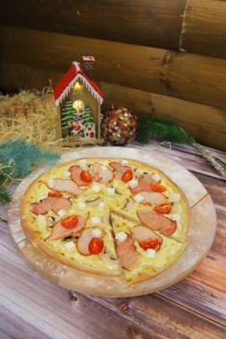 Ahşap tabakta servis edilen İtalyan etli pizza ve arka planda Noel süsü. Füme tavuk ve pastırma dilimli, feta peynirli, şampiyon mantarlı, vişneli domatesli düşey yemek.