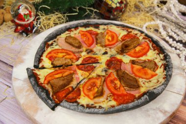 Ahşap tabakta servis edilen İtalyan siyah hamurlu pizza ve arka planda yılbaşı süsü olan masa. Domuz eti, sığır eti, domates dilimleri ve mozzarella peynirli fast food et yemeği. 