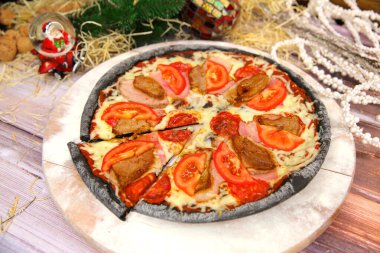 Ahşap tabakta servis edilen İtalyan siyah hamurlu pizza ve arka planda yılbaşı süsü olan masa. Domuz eti, sığır eti, domates dilimleri ve mozzarella peynirli fast food et yemeği. 
