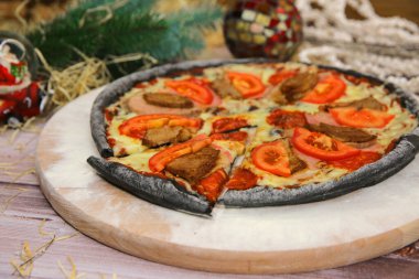 Tahta tabakta servis edilen İtalyan siyah hamurlu pizzası ve arka planda yılbaşı süslemesi. Domuz eti, sığır eti, domates dilimleri ve mozzarella peynirli fast food et yemeği. 
