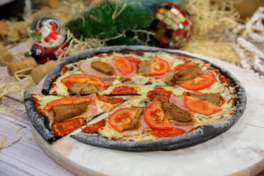 Ahşap tabakta servis edilen İtalyan siyah hamurlu pizza ve arka planda yılbaşı süsü olan masa. Domuz eti, sığır eti, domates dilimleri ve mozzarella peynirli fast food et yemeği. 