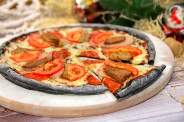 Ahşap tahtada servis edilen İtalyan siyah hamurlu pizzası ve arka planda yılbaşı süsü olan masa. Domuz eti, sığır eti, domates dilimleri ve mozzarella peynirli fast food et yemeği. 