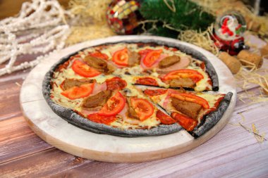 Ahşap tabakta servis edilen İtalyan siyah hamurlu pizza ve arka planda yılbaşı süsü olan masa. Domuz eti, sığır eti, domates dilimleri ve mozzarella peynirli fast food et yemeği. 