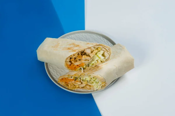 鶏肉野菜とシャワルマロール キャベツ キュウリ 薄いラバシュパンに包まれたニンジンは 薄い上にプレート上で提供 濃い青と白の抽象的な背景 ストリートフード焼きサンドイッチ — ストック写真