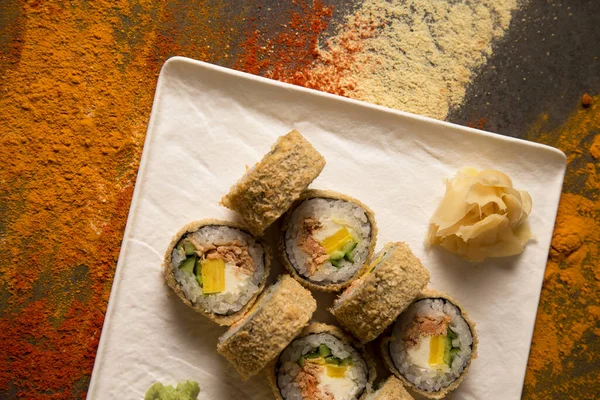 Tempura usulü sıcak suşi rulosu, kızarmış somon, krem peynir, salatalık, Tamagoyaki, zencefilli omlet, wasabi tabağında. Baharat tozunun arkaplanı: zerdeçal, köri, paprika