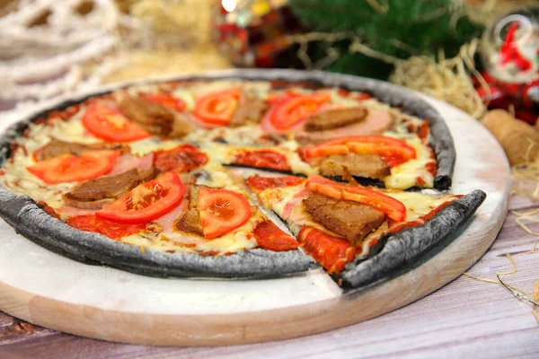 Ahşap tahtada servis edilen İtalyan siyah hamurlu pizzası ve arka planda yılbaşı süsü olan masa. Domuz eti, sığır eti, domates dilimleri ve mozzarella peynirli fast food et yemeği. 