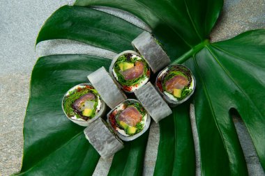 Sağlıklı Japonların hemen üstünde çiğ somon, ton balığı, kaya balığı, marul sarılı pirinç suşi rulosu yok. Gri taşlı arka planda egzotik canavar yaprağına servis ediliyor. yiyecek sanatı konsepti