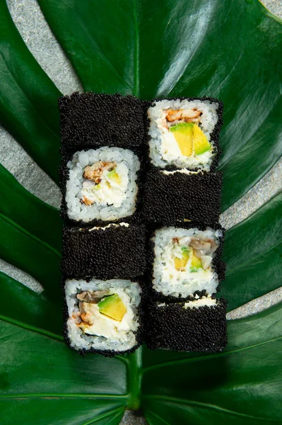 Directly above of Japanese sushi roll with unagi eel, cream cheese, avocado wrapped in black caviar (flying fish roe Tobiko) served on exotic monstera leaf on gray stone background. food art concept
