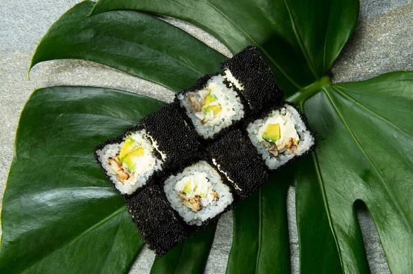 From above of Japanese sushi roll with unagi eel, cream cheese, avocado wrapped in black caviar (flying fish roe Tobiko) served on exotic monstera leaf on gray stone background. food art concept