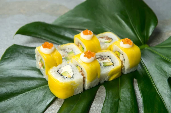 Side view of Japanese sushi roll with unagi eel, cream cheese, avocado wrapped in mango slices served with red caviar on top. exotic monstera leaf on gray stone background. food art concept