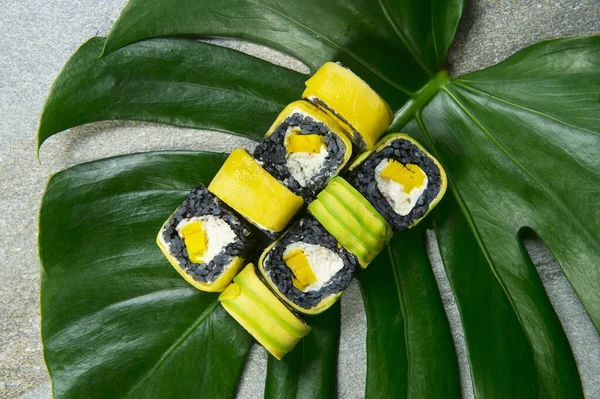 Top view of Japanese black rice sushi roll with raw eel, cream cheese, Tamagoyaki egg omelette wrapped in mango, avocado slices served on exotic monstera leaf on gray stone background. food art