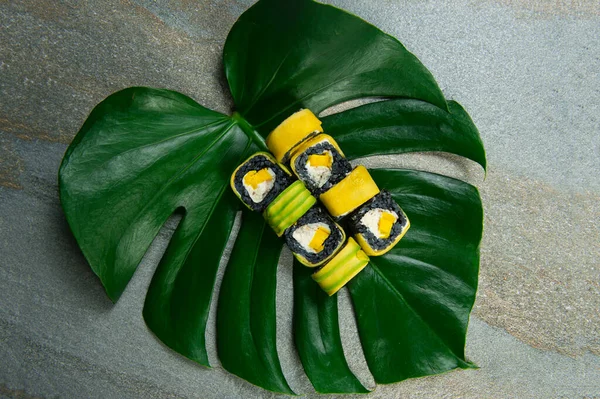 Directly above of Japanese black rice sushi roll with raw eel, cream cheese, Tamagoyaki egg omelette wrapped in mango, avocado slices served on exotic monstera leaf on gray stone background. food art