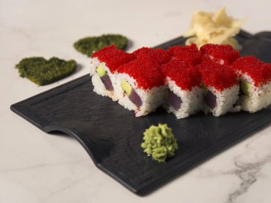 Ton balıklı Japon suşi rulosu, pirinç sarısı avokado ve üstünde kırmızı uçan balık yumurtası Tobiko. Wasabi ile servis edilen siyah tabak, zencefil. Kurumuş otlardan oluşan iki kalp mermer arka planda baharatlar.