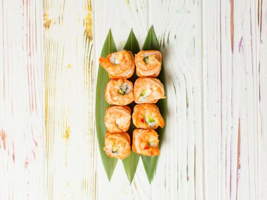 Lezzetli Japon deniz ürünleri suşi rulosu somon tataki ve üzerine kavrulmuş siyah kaplan karidesleri açık renkli ahşap arka planda bambu yaprakları ile servis edilir.