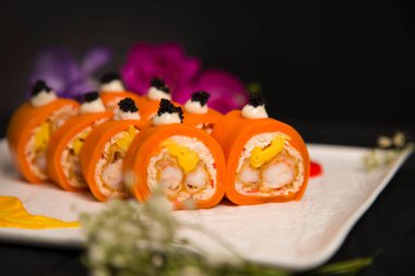 Sağlıklı Japon pirinçsiz suşi rulosunun yanında soslu havuç ve siyah havyarla servis edilen havuç karidesine sarılmış tempura siyah kaplan karidesi de var. Çiçek, fırça darbesi beyaz tabakta