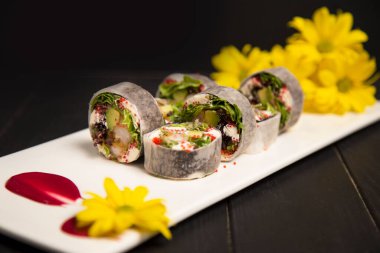 Daikon turpuna sarılmış tempura karidesli sağlıklı Japon pirinçsiz suşi rulosu. Sarı papatya çiçekleri, kırmızı boya fırça darbesi süslemesi beyaz tabakta. Kara Ahşap arkaplan