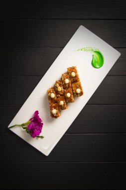 Çıtır çıtır Japon suşi rulosu sosla servis edilir. Mor çiçek çiçekleri ve dikdörtgen beyaz tabakta yeşil boya fırça darbesi süslemesi. Tam üstten. Kara tahta arka plan. Üst görünüm