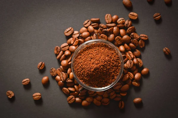 Bir avuç kahve çekirdeğinin üzerinde cam kâsede servis edilen öğütülmüş kahve. Yüksek kalitede organik Arap tohumları. Koyu arkaplana saçılmış kahve çekirdekleri odak dışı. Boşluk metin resmini kopyala