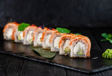 Japon suşisi tahta zemin üzerinde siyah taş tabakta yuvarlanıyor. Philadelphia krem peynirli suşi parçaları, pirinçle sarılmış avokado üzerine yanmış somon Tataki fileto ve yeşillik.