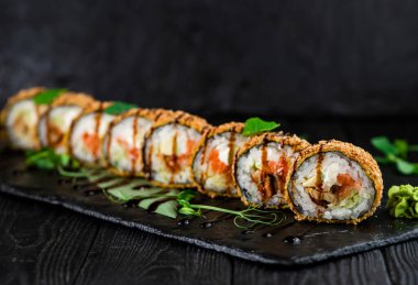 Japon tempura sıcak suşi rulosu tahtadan arka planda siyah taş plaka üzerinde. Suşi parçaları somon, yılan balığı, salatalık, krem peynir, pirinç sarılı avokado ve sebzeli gevrek yosun.