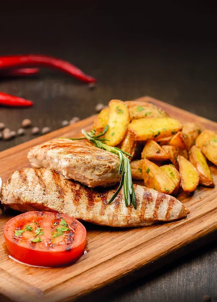 Vertical image shot of Grilled Chicken steak on wooden board served with grilled tomato slice and homemade baked potatoes. Garlic sauce in white bowl. Rosemary brunch on meat loaf.