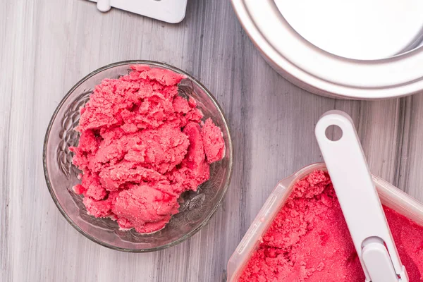 homemade cherry ice cream in a plastic container on the table next to an ice cream spoon and an ice cream maker