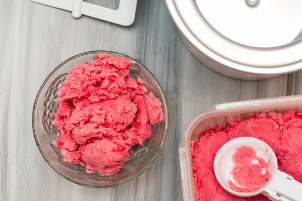 homemade cherry ice cream in a plastic container on the table next to an ice cream spoon and an ice cream maker