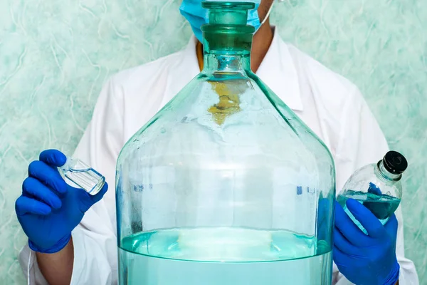 Um médico no laboratório tem frascos de líquido nas mãos há outro grande recipiente na mesa — Fotografia de Stock