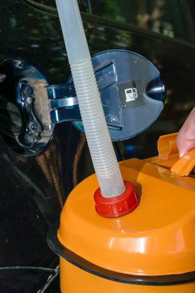 Mężczyzna trzyma pomarańczowy kanister z benzyną w ręku obok samochodu z wężem odpływowym przykręconym do szyi właz zbiornika paliwa jest otwarty. — Zdjęcie stockowe