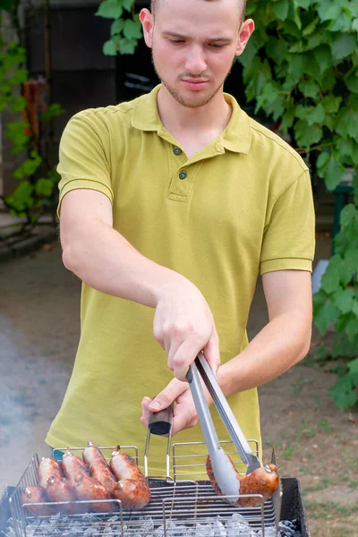 Genç bir adam bahçedeki kömürlerin üstünde ızgarada sosis pişiriyor. Yüksek kalite fotoğraf