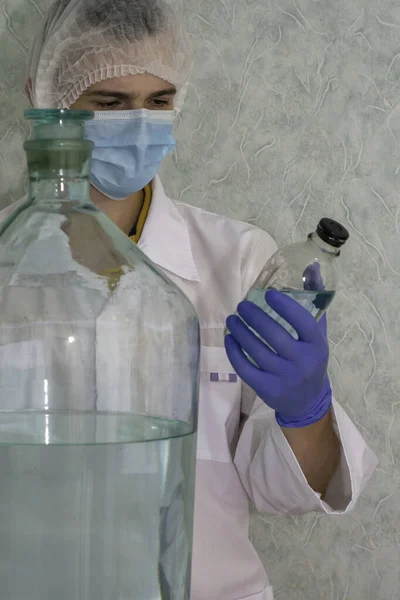a medical laboratory assistant in a white coat cap gloves and a protective mask examines a glass bottle with a liquid holding it in his hand there is a huge vessel with a liquid next to it concept vaccination pharmaceuticals. High quality photo