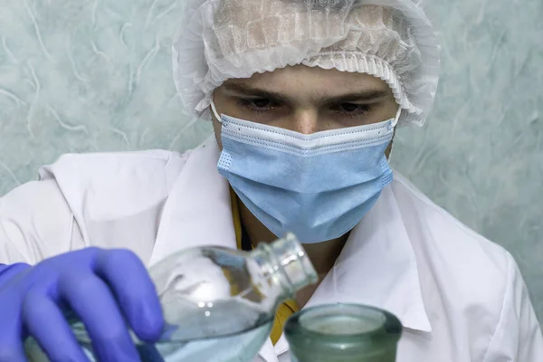 a medical laboratory assistant in a white coat gloves a hygienic mask and a cap pours liquid from one vessel to another of a larger volume. High quality photo