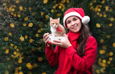 Kırmızı Noel Baba şapkalı sevimli Avrupalı bir kız hediye olarak küçük kırmızı bir kedi yavrusu alıyor ve mutlu oluyor. Hediye olarak bir evcil hayvan al ve kutusundan çıkar. Sarı çelenklerle dolu parlak yeni yıl arkaplanı.